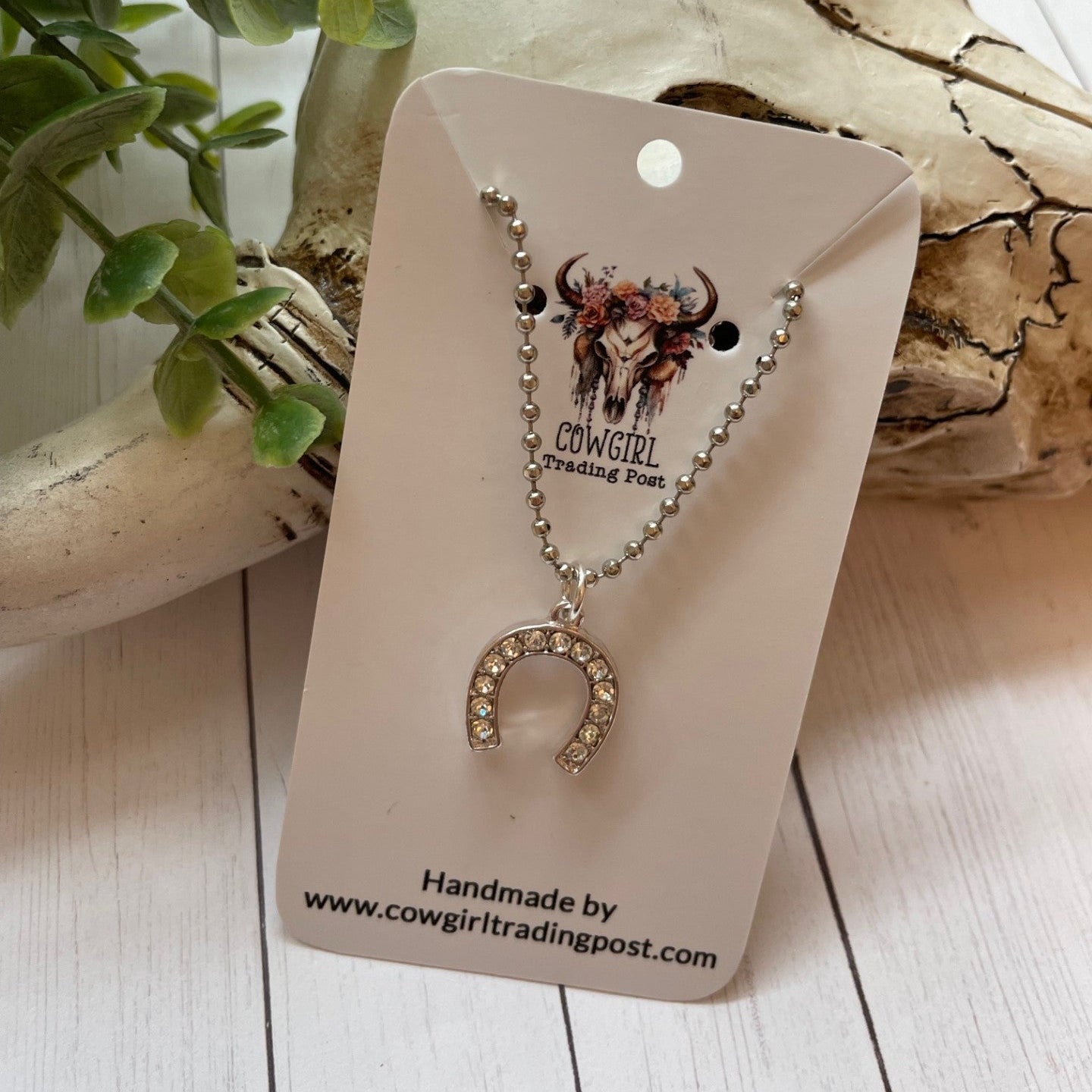 Necklace with horseshoe pendant on a 'Cowgirl Trading Post' card, surrounded by natural elements.