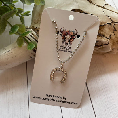 Necklace with horseshoe pendant on a 'Cowgirl Trading Post' card, surrounded by natural elements.