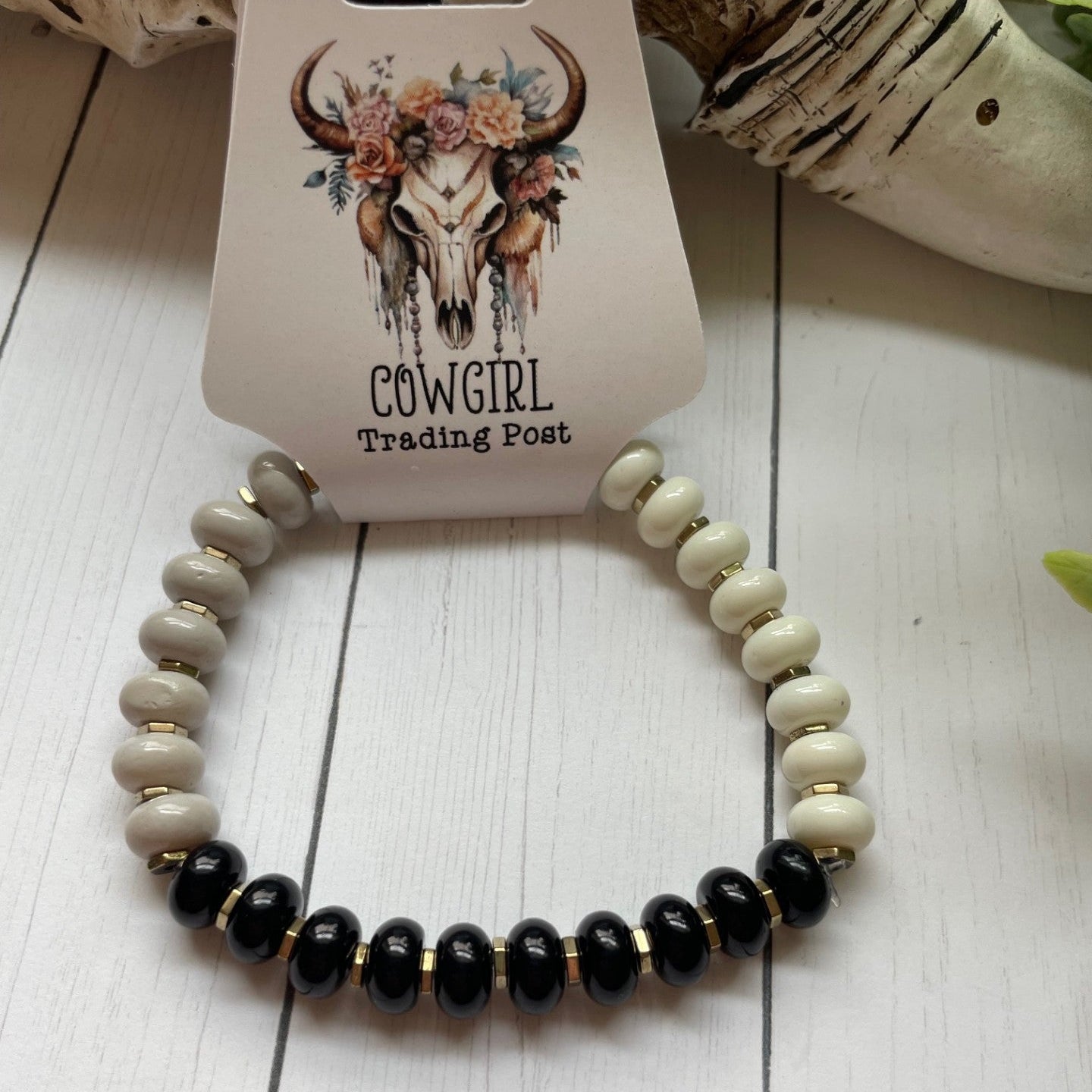 A beaded stretch bracelet displayed on its branded card. The bracelet features alternating sections of glossy black beads and matte cream/off-white beads, separated by small gold spacer rings.