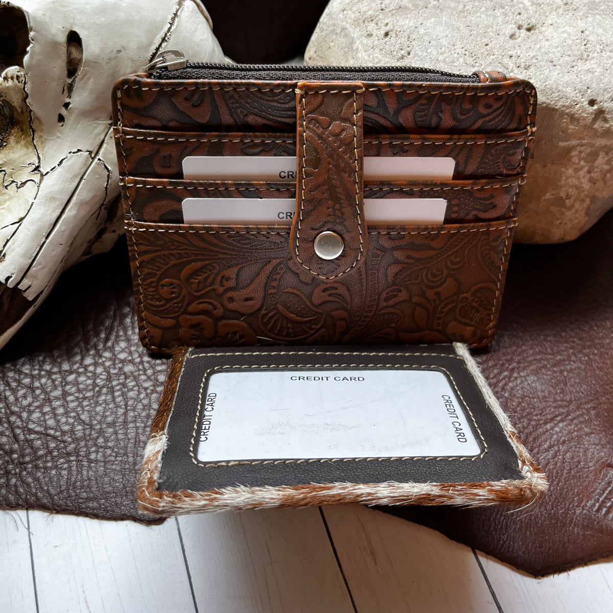 A brown embossed leather cardholder wallet with a snap-button closure, shown open with cards inserted, placed on a dark leather surface with a credit card holder and decorative stones nearby.