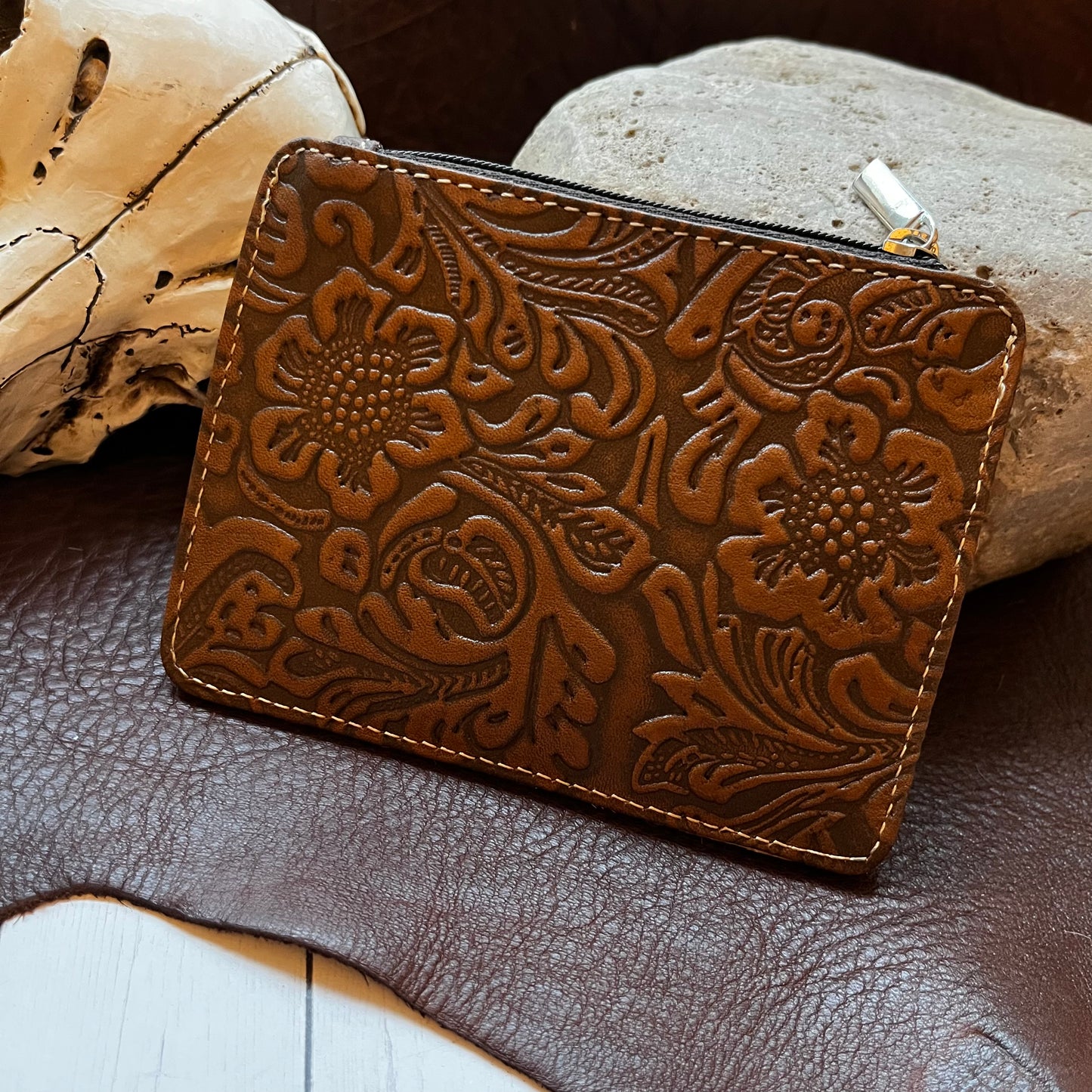 A brown embossed leather zip-around pouch with floral tooling, styled on a dark leather surface with a skull and rock props.