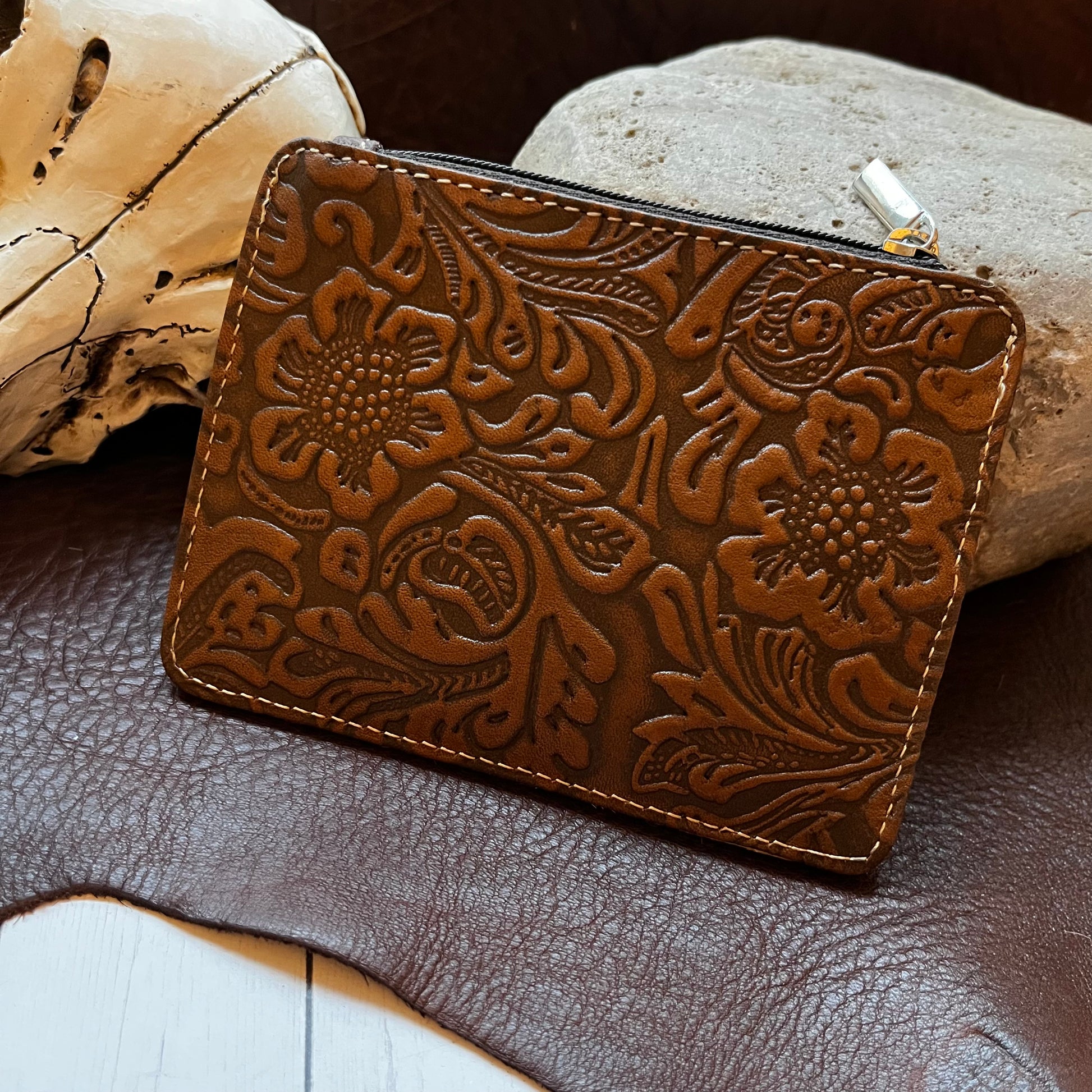 A brown embossed leather zip-around pouch with floral tooling, styled on a dark leather surface with a skull and rock props.