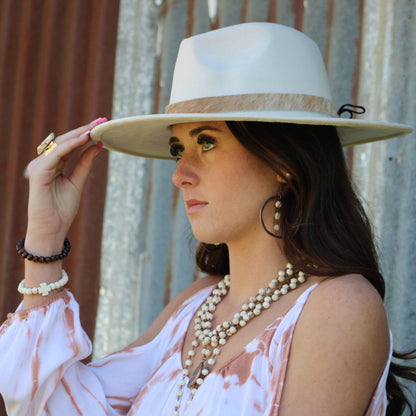 Woman wearing a wide-brimmed hat and multiple necklaces against a rustic background