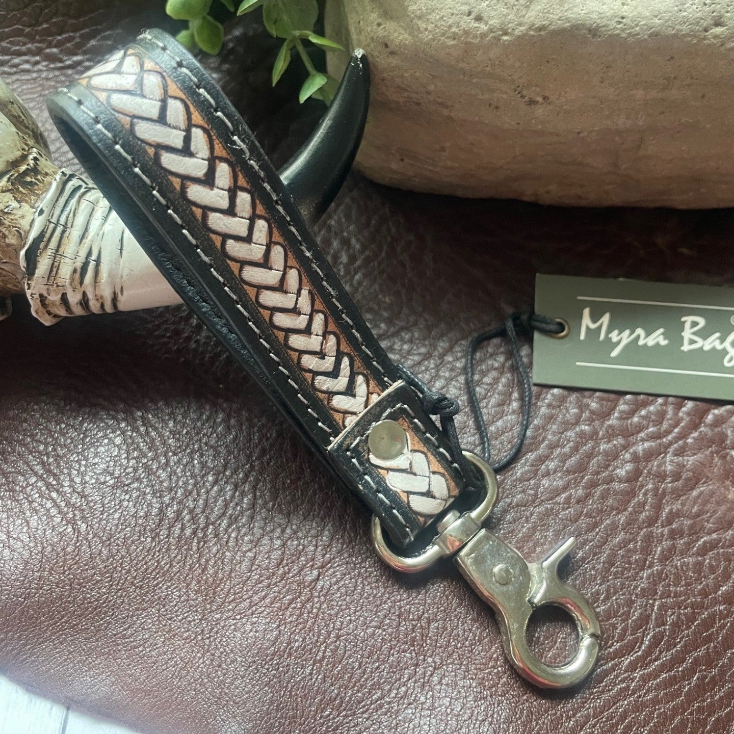 A close-up of a braided leather strap or keychain accessory with a silver metal clasp, resting on a brown textured leather surface next to a plant, stone, and a small tag reading 'Myra Bag'.
