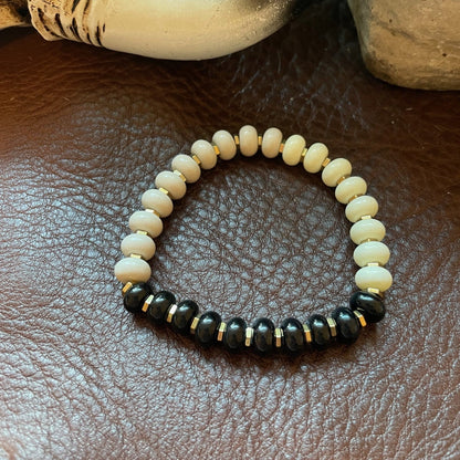 A beaded stretch bracelet with cream-colored round beads and a front section of glossy black faceted beads separated by small gold spacers, displayed on a brown textured leather surface with decorative props in the background.