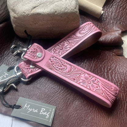 A pink tooled leather keychain or bag charm with floral embossing, shown with metal hardware and a branded paper tag reading Myra Bag, placed on a brown leather surface with stones in the background.
