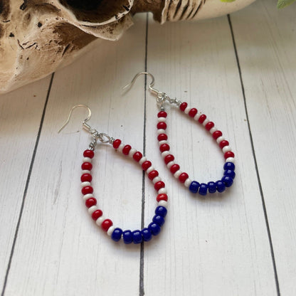 red white and blue teardrop shaped earrings in a teardrop shape on a white card with white wood background and a skull and greenery