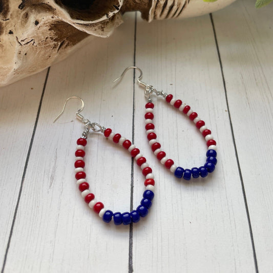 red white and blue teardrop shaped earrings in a teardrop shape on a white card with white wood background and a skull and greenery