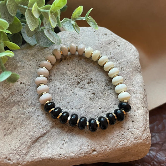 A beaded stretch bracelet with alternating black and cream beads, accented by small gold spacer rings, displayed on a textured stone with green leaves in the background.