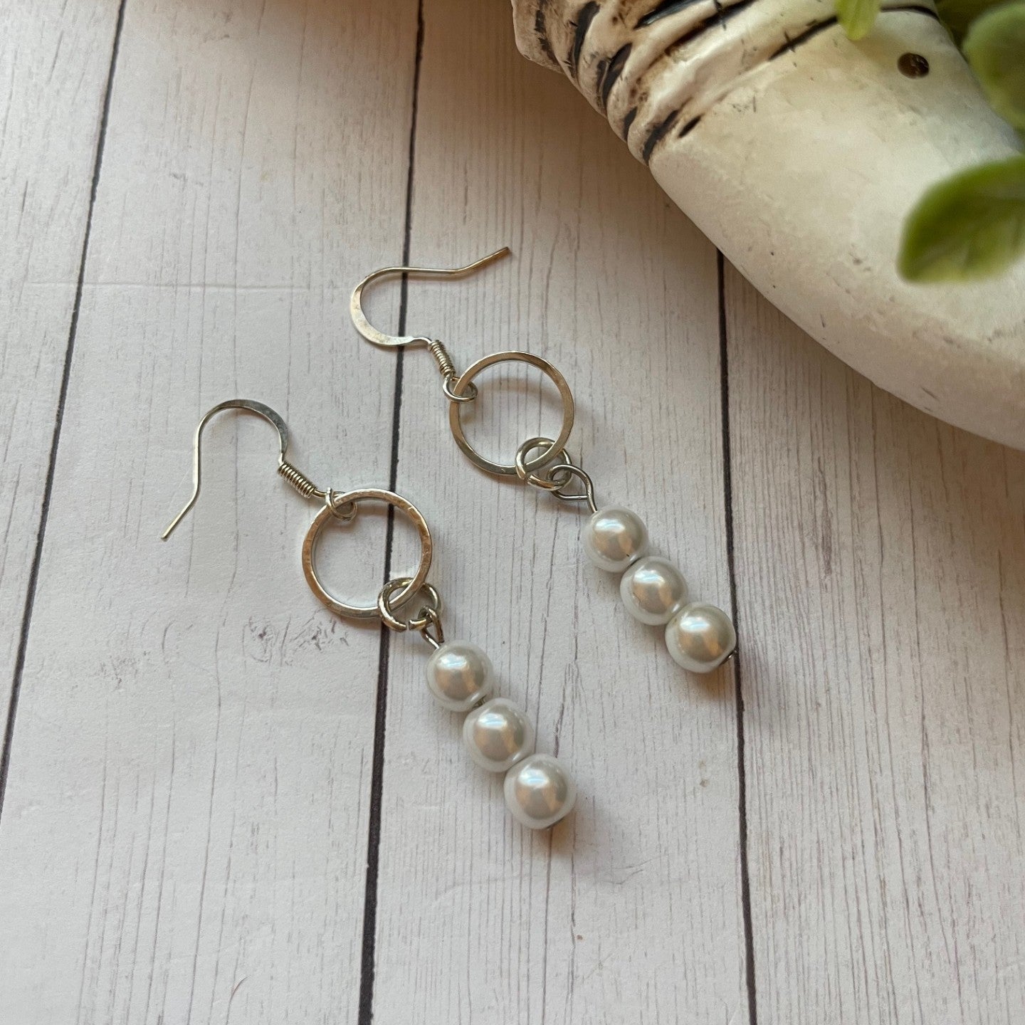 Pearl drop earrings on a wooden surface with a shell and plant in the background
