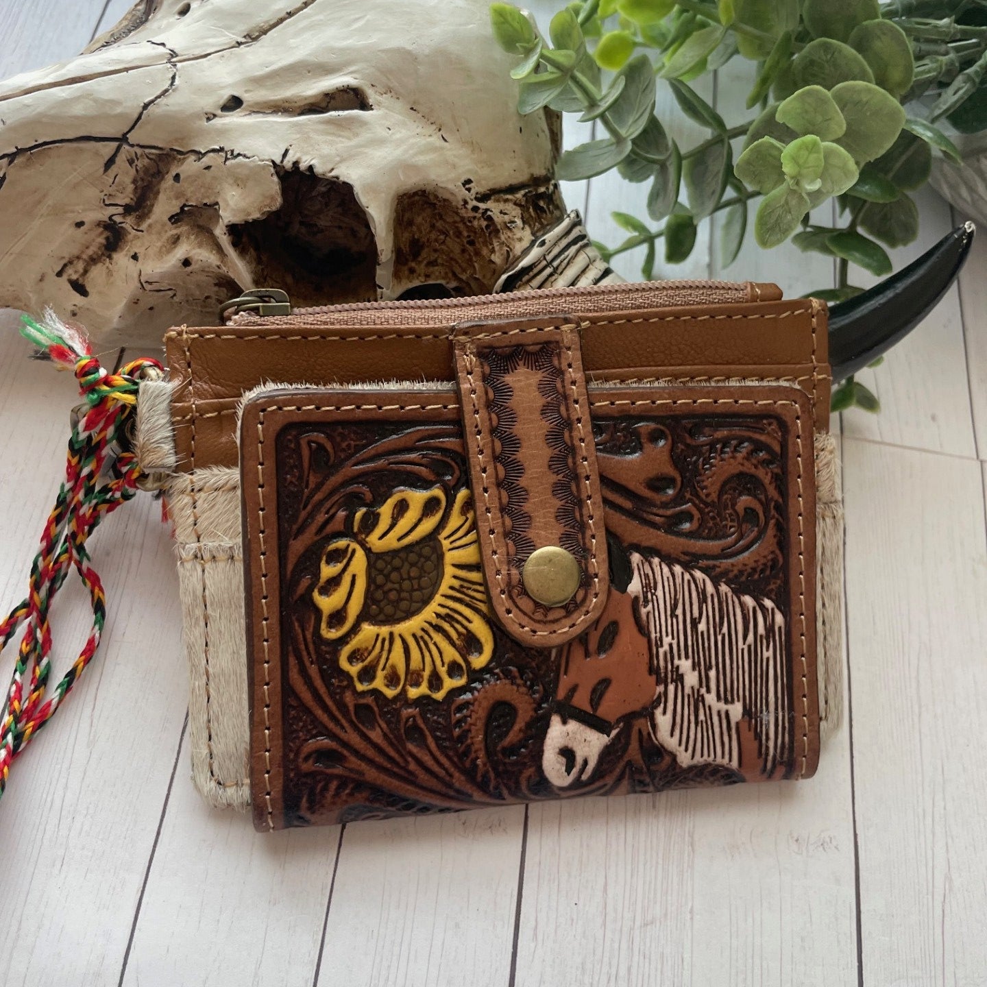 Brown leather wallet with the face head of a horse and a sunflower design on a wooden surface with plants in the background