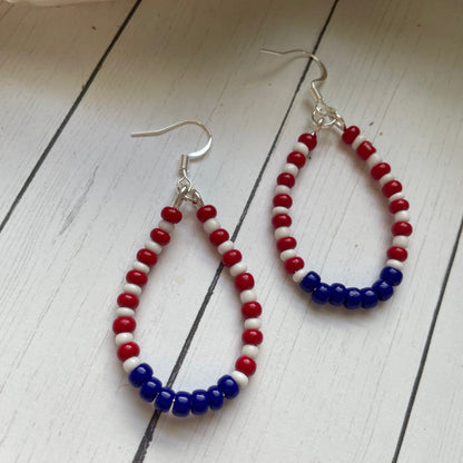 red white and blue teardrop shaped earrings in a teardrop shape on a white card with white wood background and a skull and greenery