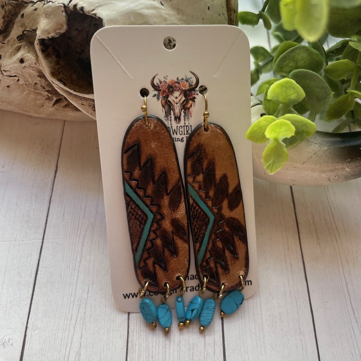 Leopard print earrings with turquoise accents on a white card, placed on a wooden surface with a plant in the background.