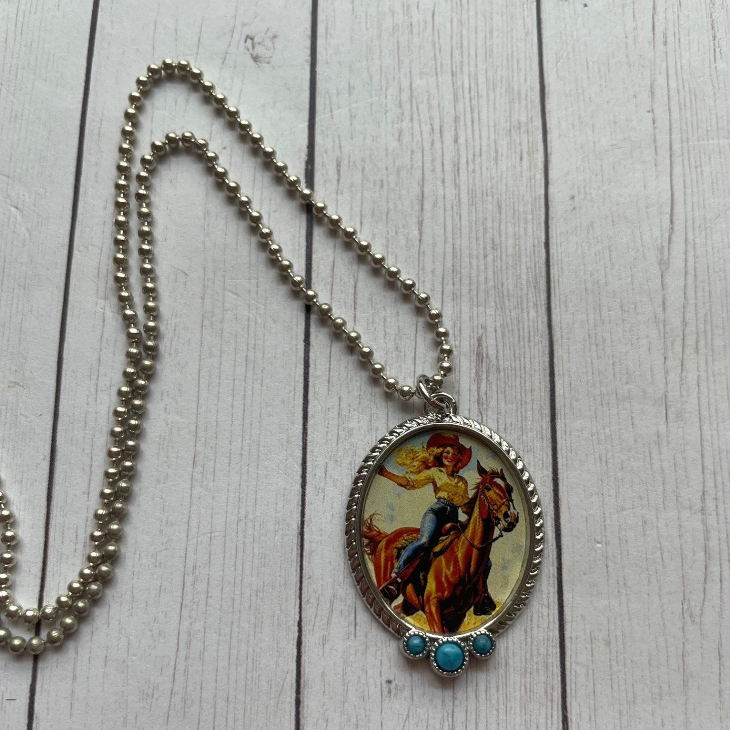 A silver beaded chain necklace with an oval pendant featuring a colorful illustration of a cowboy on a horse, resting on a white wood plank surface.