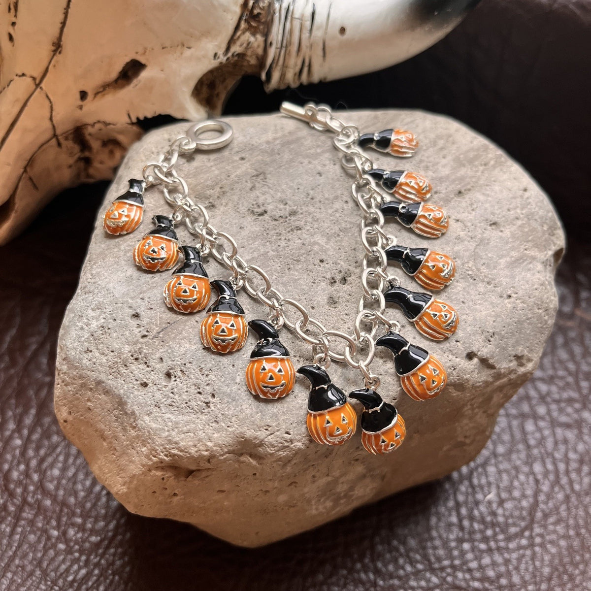 A silver chain bracelet with multiple dangling orange and black witch-hat pumpkin charms, displayed on a gray stone with a skull prop in the background.