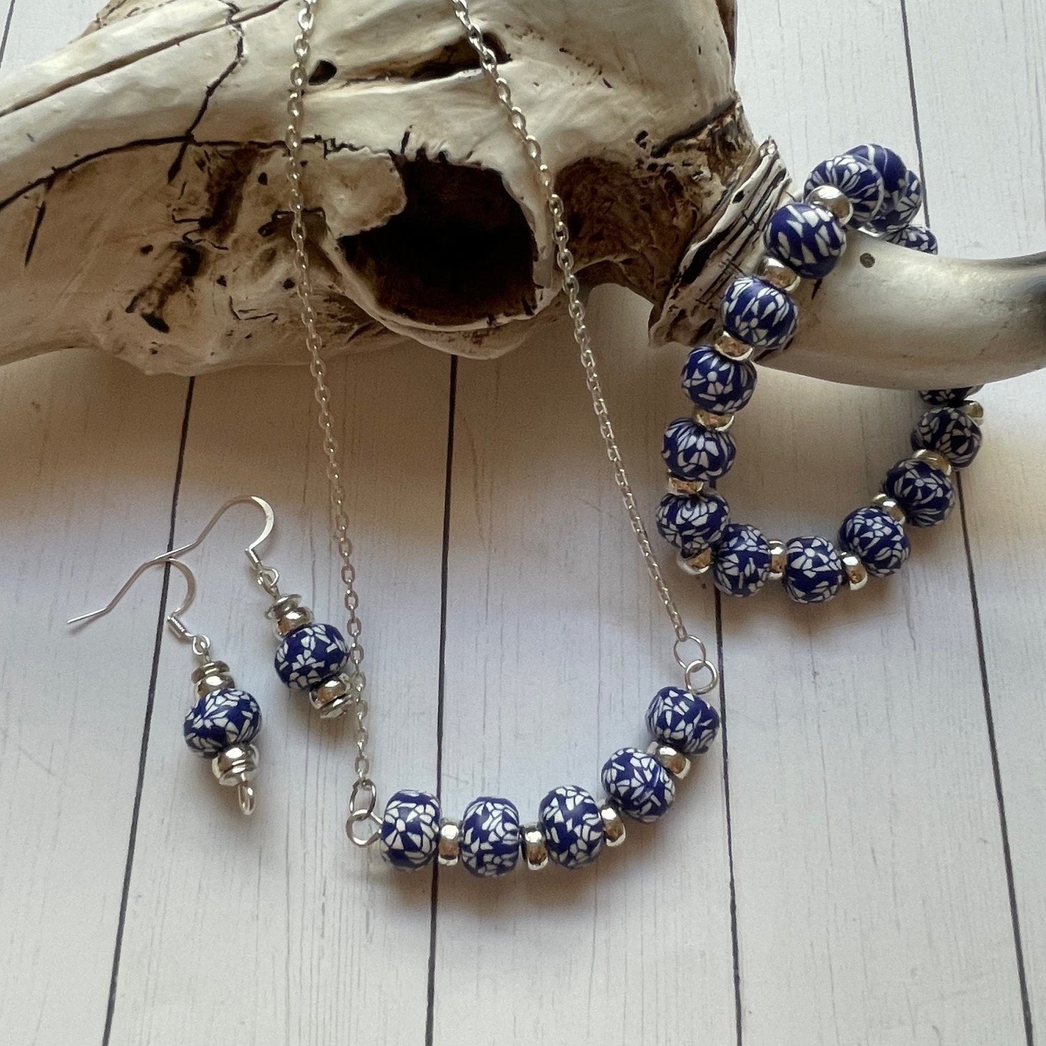 A jewelry set featuring blue and white patterned beads with silver-toned metal chain and hooks, styled on a white wood background next to a bone prop.