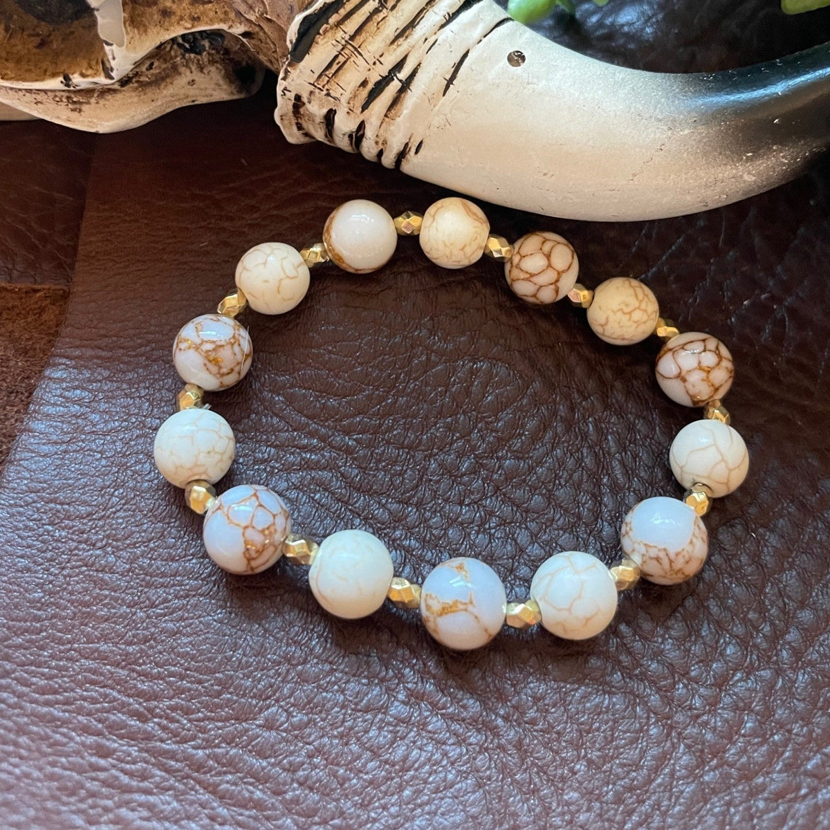 A beaded stretch bracelet made of round white and cream marbled beads with gold spacer beads, displayed on a brown leather surface next to a decorative horn and green leaves.