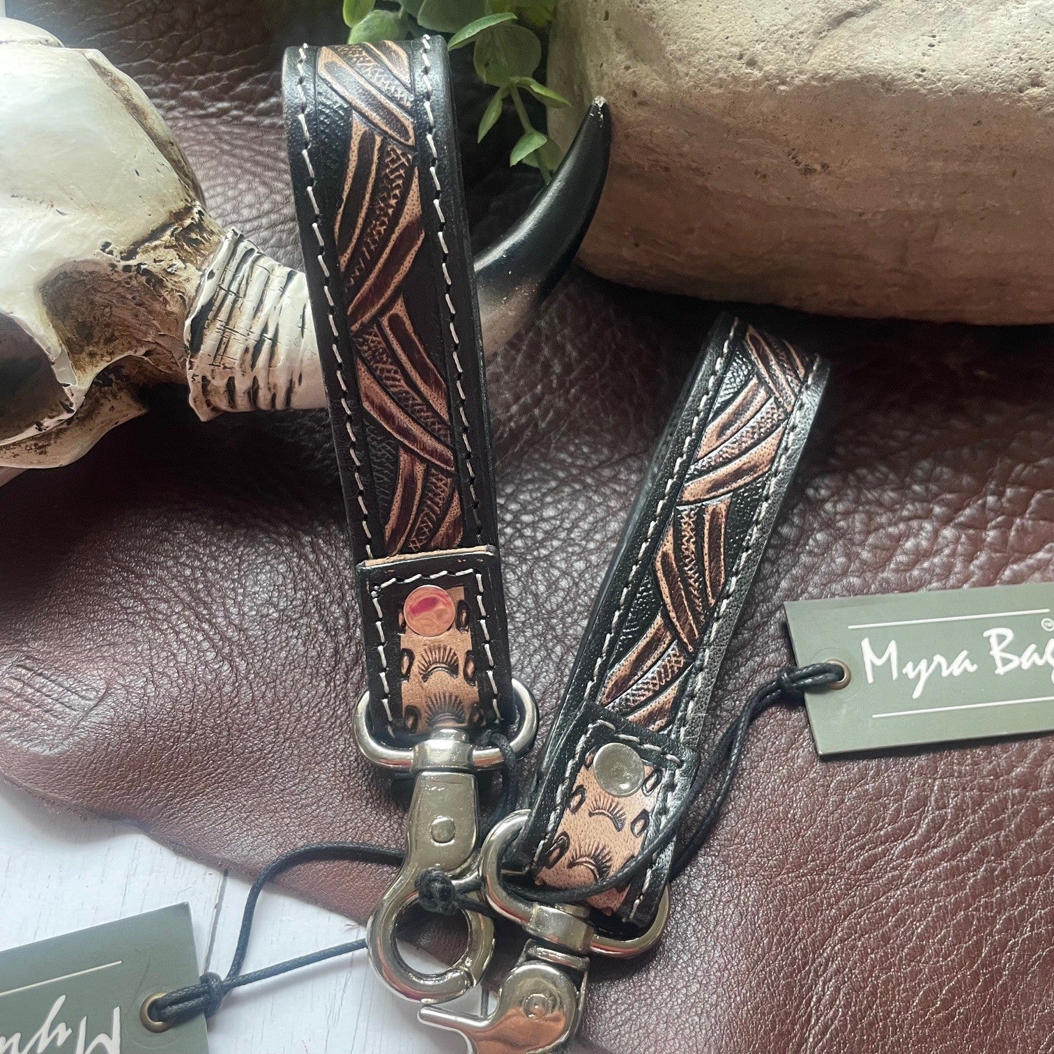 Two black leather strap accessories with embossed floral and geometric patterns in brown and bronze tones, featuring silver-tone metal clasps, displayed on a brown leather surface with decorative props and Myra Bag brand tags.