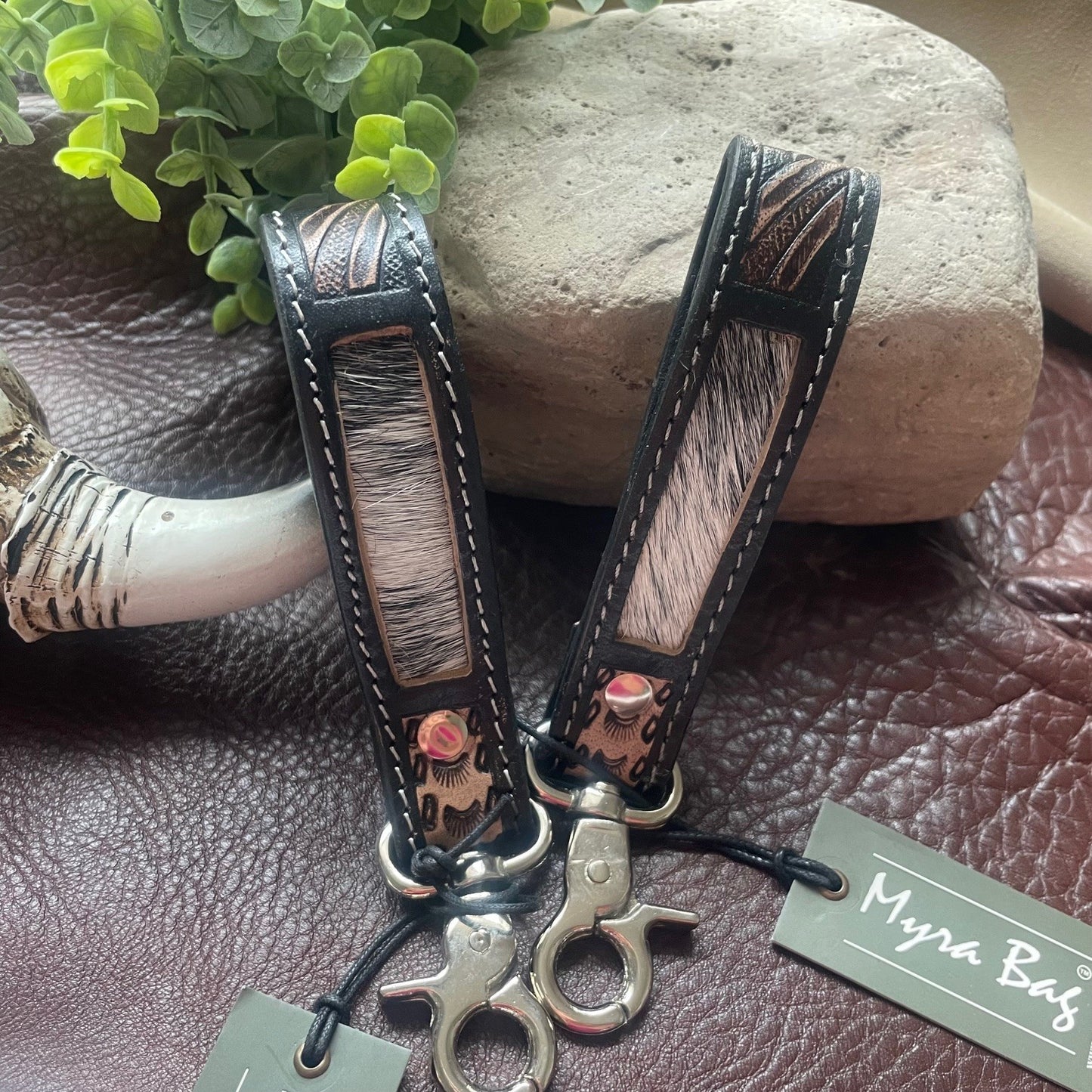 Two leather keychains with mixed animal-print and black leather panels, silver-tone hardware, and hanging tags reading 'Myra Bag', styled on a brown leather surface with a stone and green leaves in the background.