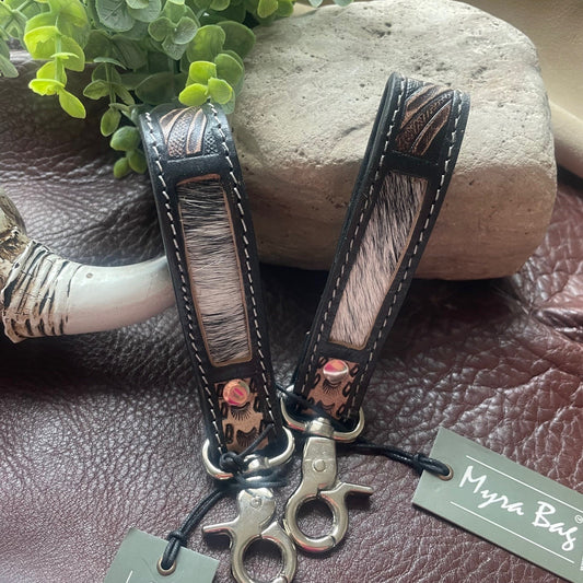Two leather keychains with mixed animal-print and black leather panels, silver-tone hardware, and hanging tags reading 'Myra Bag', styled on a brown leather surface with a stone and green leaves in the background.