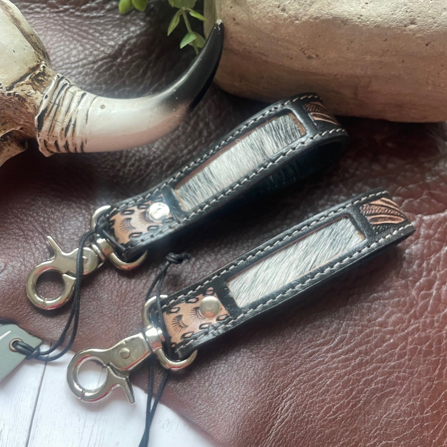 Two leather keychains with textured silver-gray hair-on-hide panels and copper-toned metal studs, displayed on a brown leather surface with rustic props.