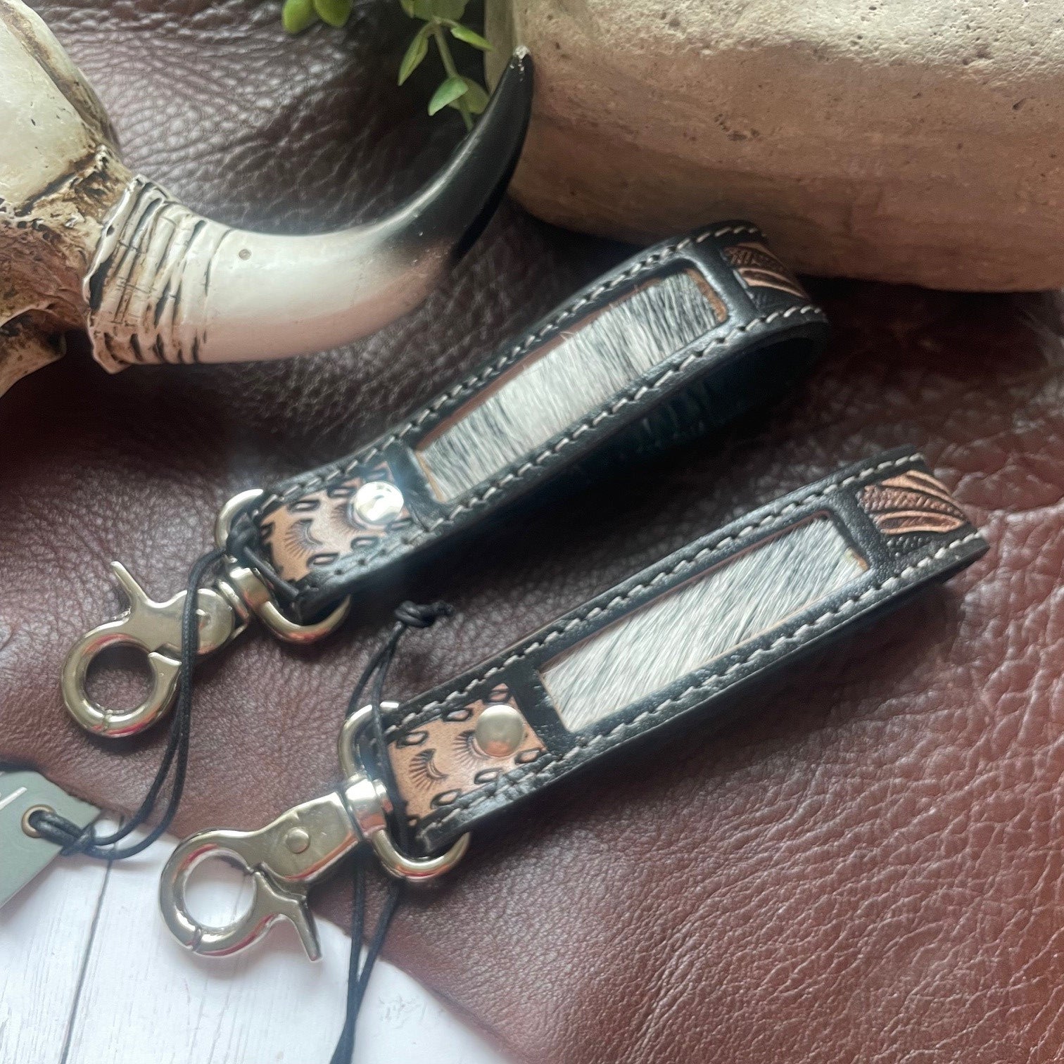 Two leather keychains with textured silver-gray hair-on-hide panels and copper-toned metal studs, displayed on a brown leather surface with rustic props.