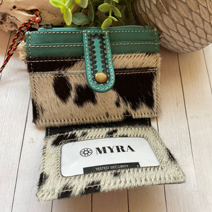 Small wallet with cow print and teal accents on a wooden surface, featuring the brand 'MYRA'.