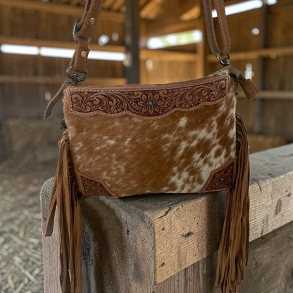 A western-style shoulder bag made of brown and white cowhide with tooled leather trim and long brown fringe, displayed on a wooden post inside a barn.