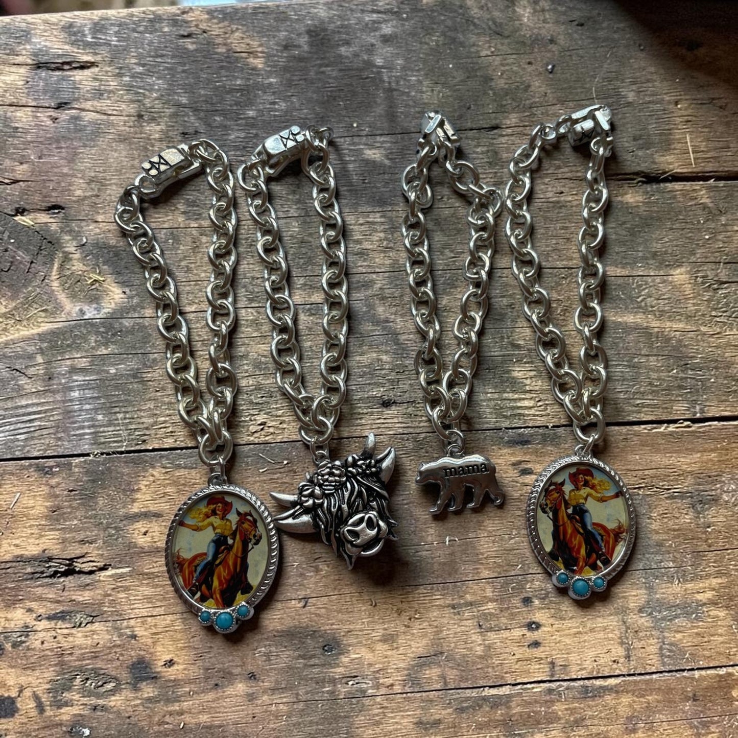 four silver chain bracelets with oval enamel pendants and sculptural animal charms, displayed on a rustic wooden surface. One pendant features a cowboy riding a horse, and the other features a silver bear with mama bear iimprinted