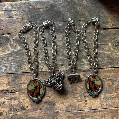 four silver chain bracelets with oval enamel pendants and sculptural animal charms, displayed on a rustic wooden surface. One pendant features a cowboy riding a horse, and the other features a silver bear with mama bear iimprinted