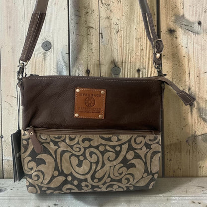 A brown leather crossbody bag with a tan and brown damask patterned lower half, featuring a brown leather brand patch that reads 'LIFE & BLOOM LAFAYETTE, KY'. The bag is shown against a rustic wooden plank background.