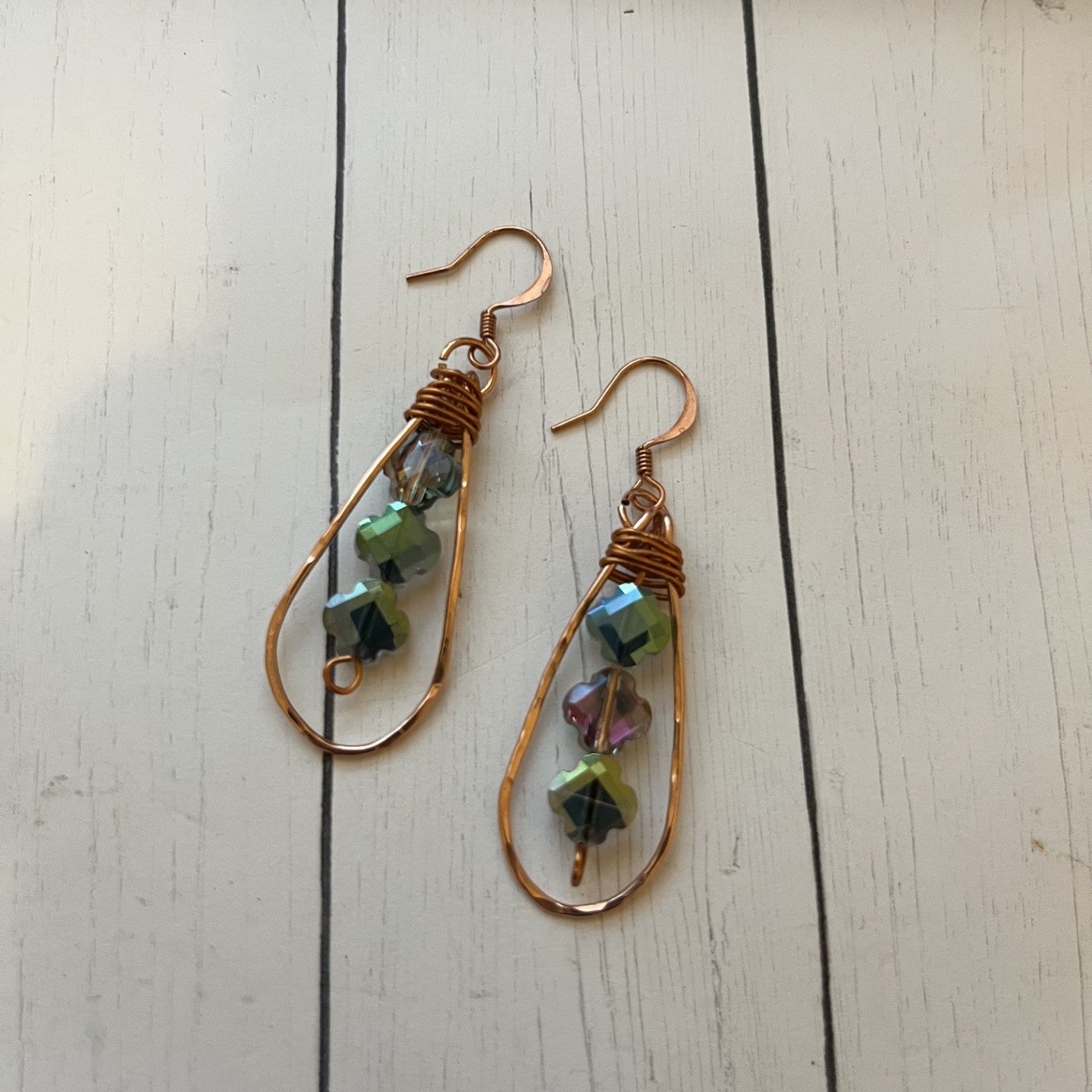 A pair of handmade teardrop dangle earrings with copper wire frames, wrapped copper wire at the top, and small faceted iridescent beads in green, blue, and pink, displayed on a white wooden surface.