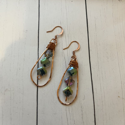 A pair of handmade teardrop dangle earrings with copper wire frames, wrapped copper wire at the top, and small faceted iridescent beads in green, blue, and pink, displayed on a white wooden surface.