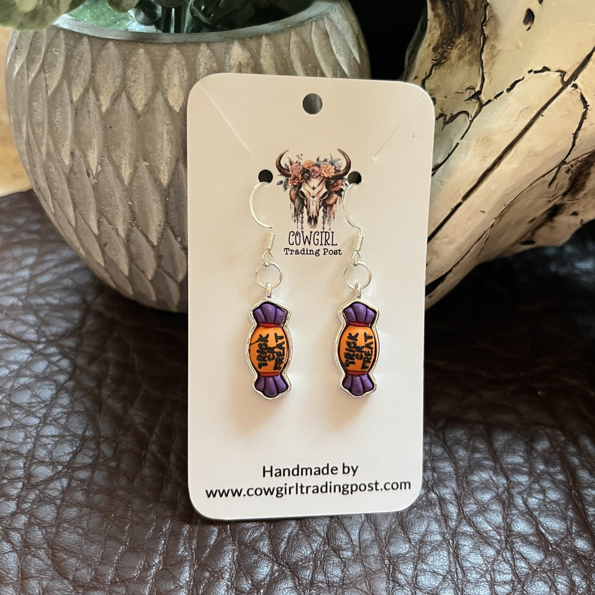 A pair of handmade dangle earrings displayed on a white backing card. The earrings feature purple wrapped candy charms with orange and black illustrated details, hanging from silver fishhook wires.