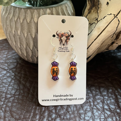 A pair of handmade dangle earrings displayed on a white backing card. The earrings feature purple wrapped candy charms with orange and black illustrated details, hanging from silver fishhook wires.