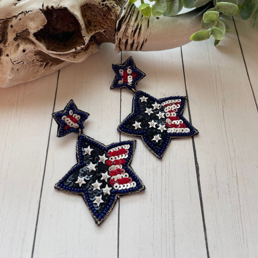 Star-shaped earrings with American flag design on a wooden surface with driftwood and plant.