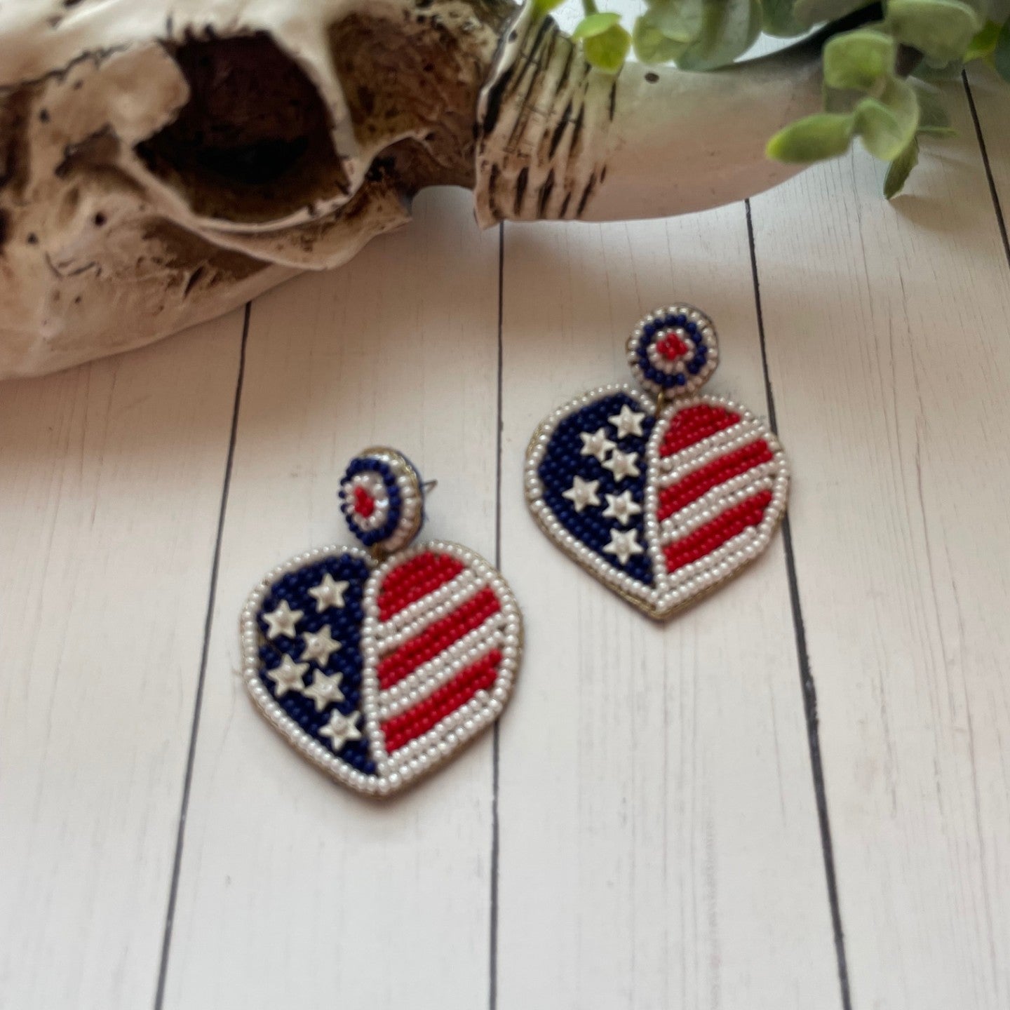 Heart-shaped earrings with American flag design on a wooden surface with a skull and plant in the background.