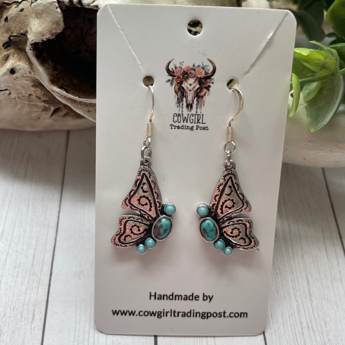 Butterfly-shaped earrings with turquoise stones on a Cowgirl Trading Post card, set against a wooden surface.