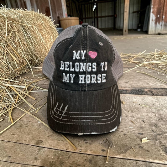 Black and gray cap with text 'My heart belongs to my horse' on a wooden floor with hay.