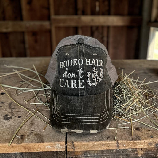 Black and gray cap with 'Rodeo Hair Don't Care' text on a wooden surface with hay.