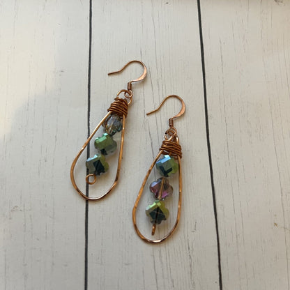 A pair of handmade teardrop dangle earrings with copper wire frames, wrapped copper wire at the top, and small faceted iridescent beads in green, blue, and pink, displayed on a white wooden surface.