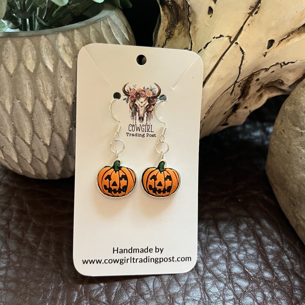 A pair of orange jack-o'-lantern dangle earrings with green stems and black facial details, hanging from silver hooks on a white display card. The card is photographed on a brown textured surface with decorative stone and wood props in the background.