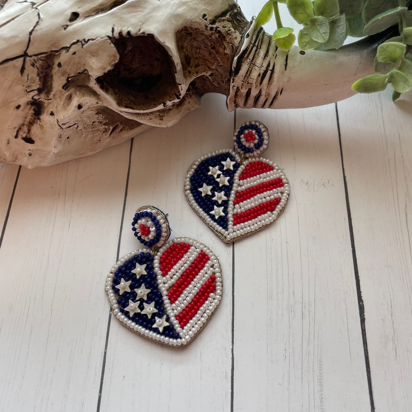 Heart-shaped patriotic earrings on a wooden surface with a skull and plant in the background