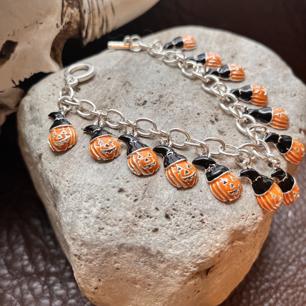 A silver chain bracelet with multiple pumpkin charms, displayed on a textured gray stone. The charms are orange and black with carved jack-o'-lantern faces.