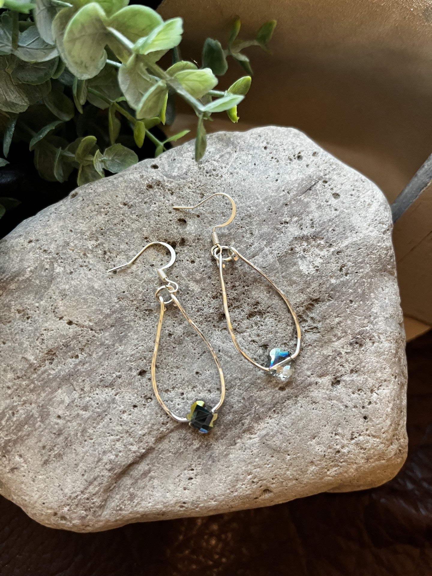 A pair of handmade teardrop dangle earrings with gold wire frames, small clear beads, and dark blue-green stone accents, displayed on a textured stone surface with green foliage in the background.