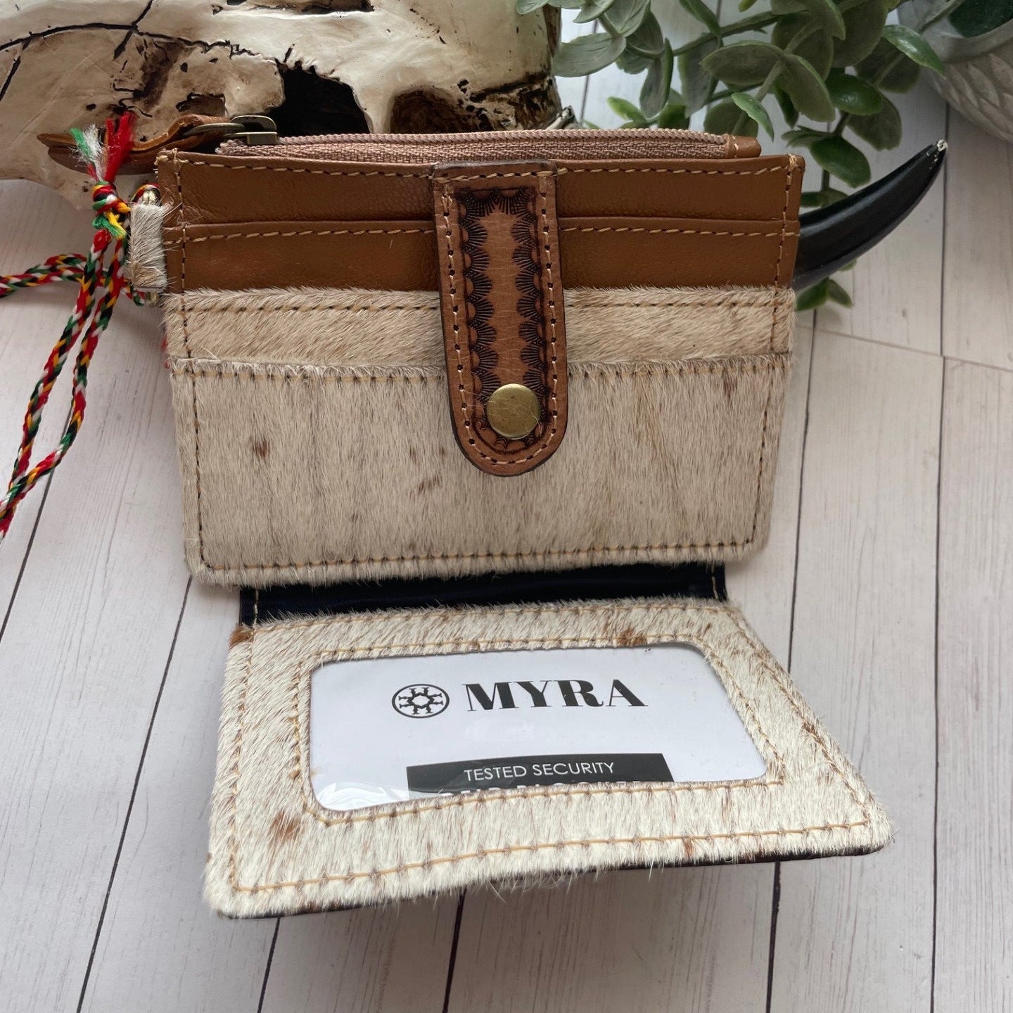Brown leather wallet with brown and white cowhide and credit card slots on a wooden surface with plants in the background