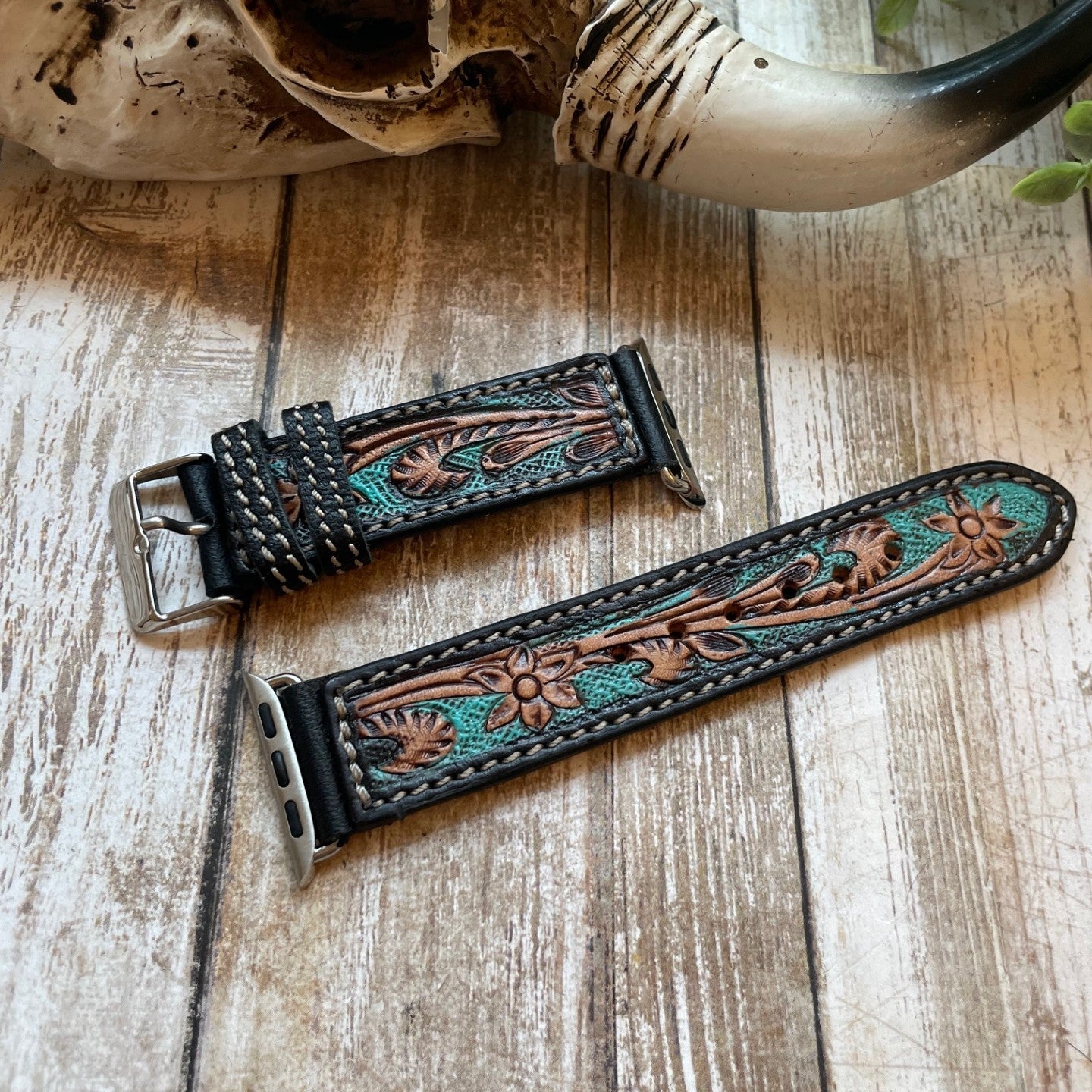 Two intricately designed leather watch straps on a wooden surface with a skull and horns in the background.