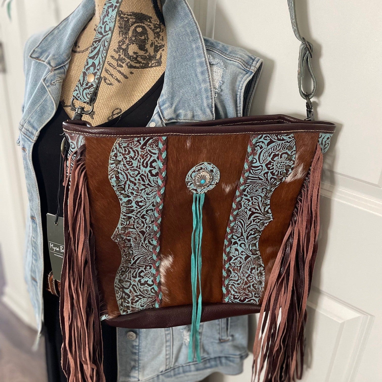 Brown leather handbag with fringe and patterned design hanging on a mannequin against a white wall.