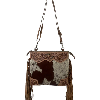 A western-style crossbody handbag featuring a brown and black cowhide patterned front, tooled leather detailing, and long brown fringe, shown against a plain white background.