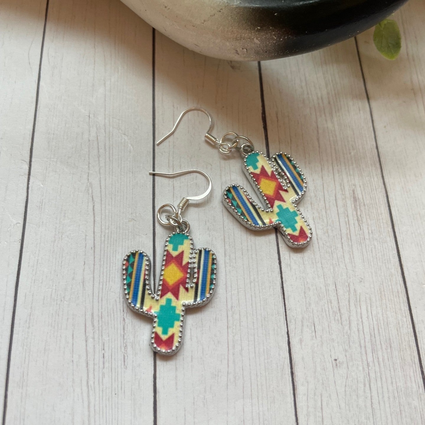Colorful cactus-shaped earrings on a wooden surface with a plant in the background